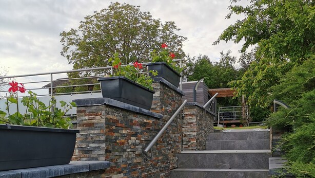 Un escalier avec des marches en pierre et des rampes en métal, menant à un espace abrité agrémenté de verdure.