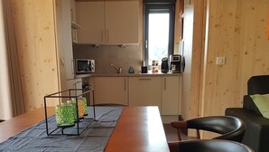 Une table de salle à manger en bois, accompagnée de quatre chaises, un coin cuisine avec des placards et des appareils électroménagers, et une fenêtre laissant entrer la lumière naturelle.