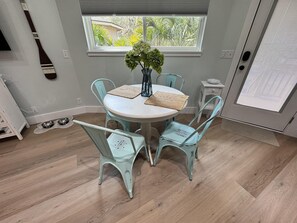 A dining area with a round table, four chairs, a vase with flowers, and a window with blinds.