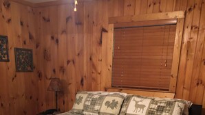 A wooden-paneled room with a large bed, a nightstand, a lamp, and a window with blinds.