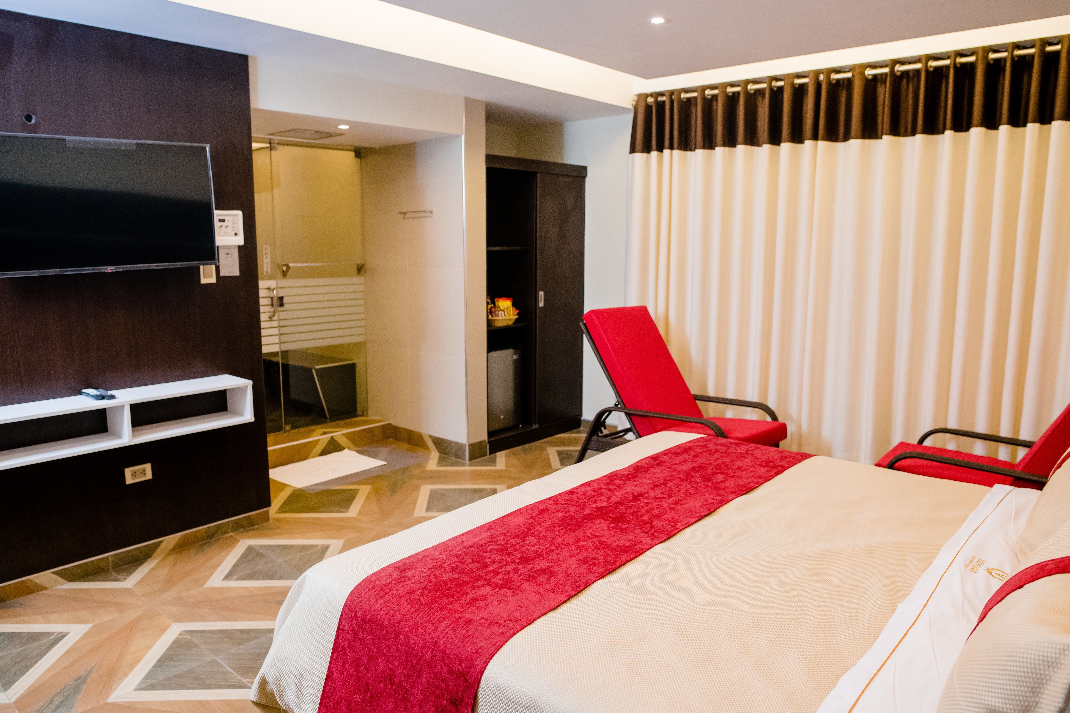 Habitación de hotel con cama, televisión montada en la pared, una silla roja y baño visible a través de una puerta abierta.