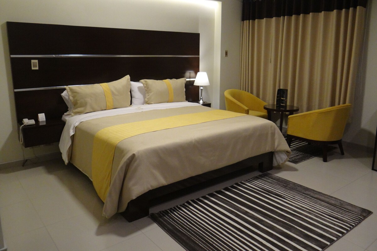 A hotel room with a large bed, a bedside table with a lamp, a chair, and a view of the outside through curtains.