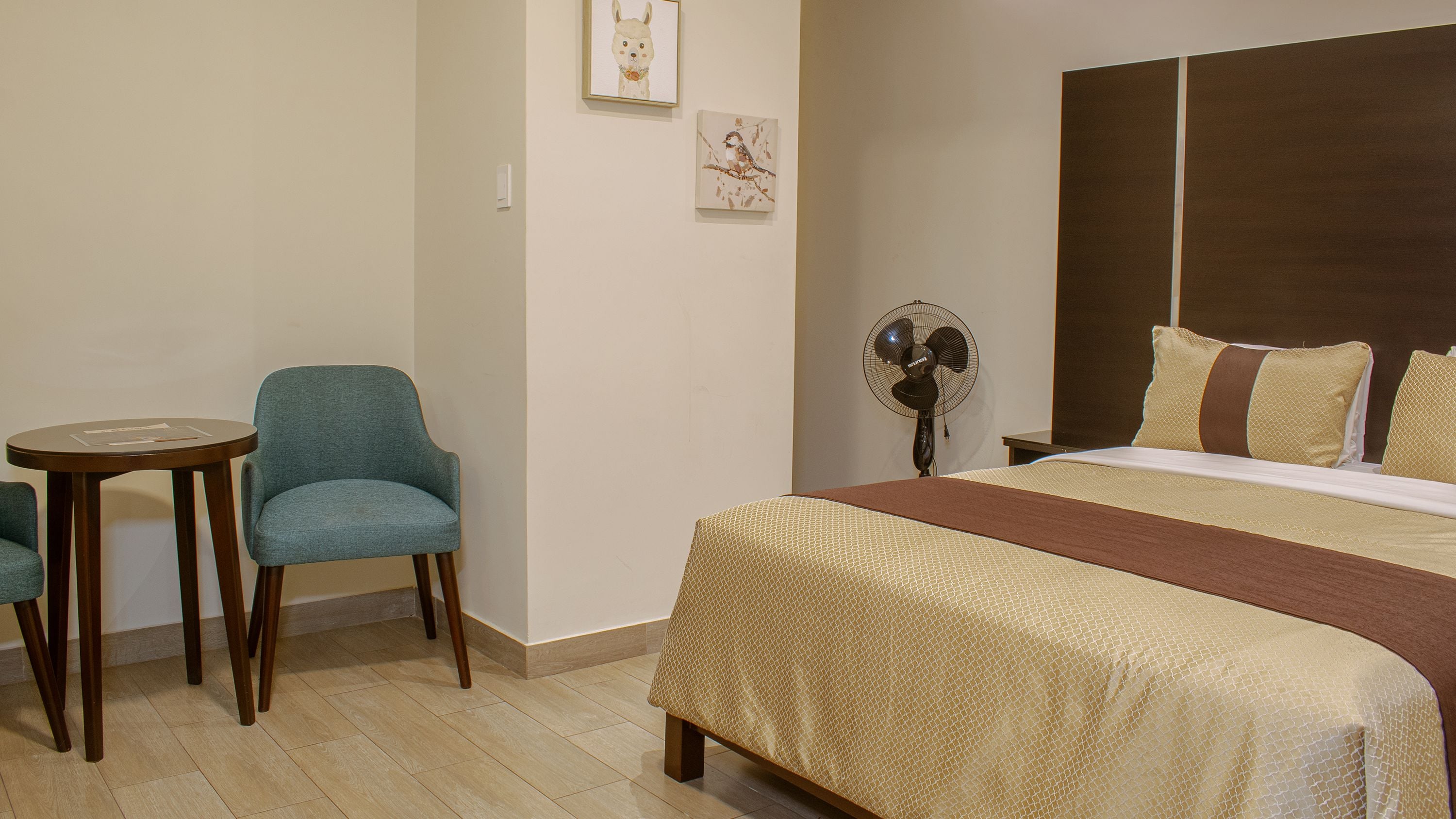Una habitación de hotel con una cama, dos sillas, una mesita pequeña y un ventilador fijado a la pared.