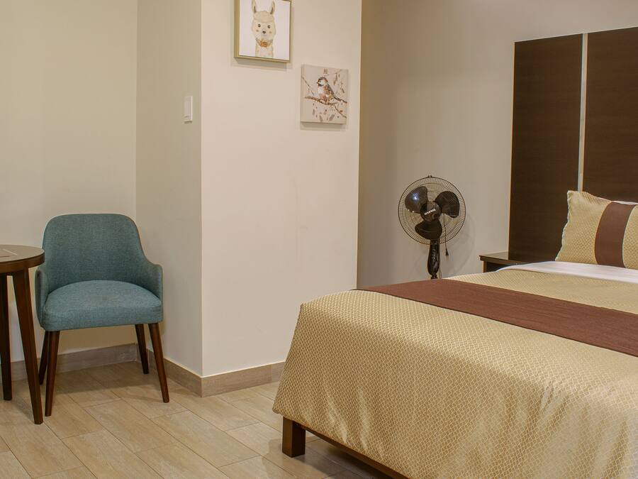 Una habitación de hotel con una cama, dos sillas, una mesita pequeña y un ventilador fijado a la pared.