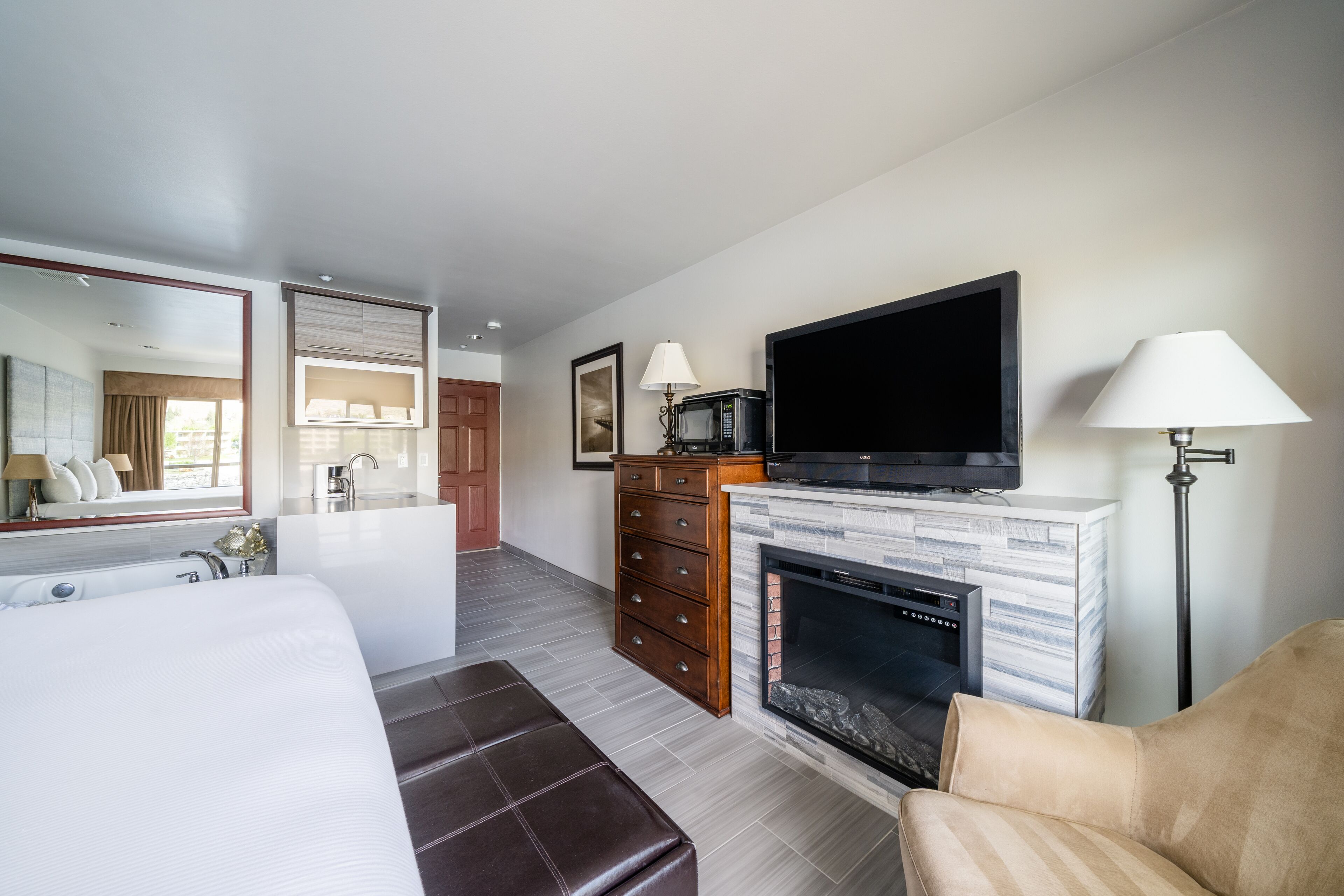 A modern hotel room with a large bed, a flat-screen TV, a fireplace, a desk, and a bathroom with a large mirror and a bathtub.
