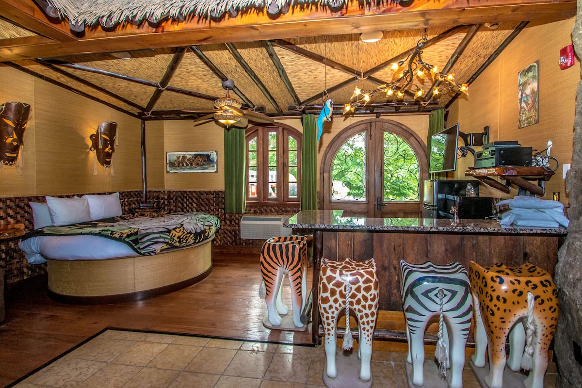 A room with a curved bed, a wooden ceiling, a bar area with stools, and a large window.