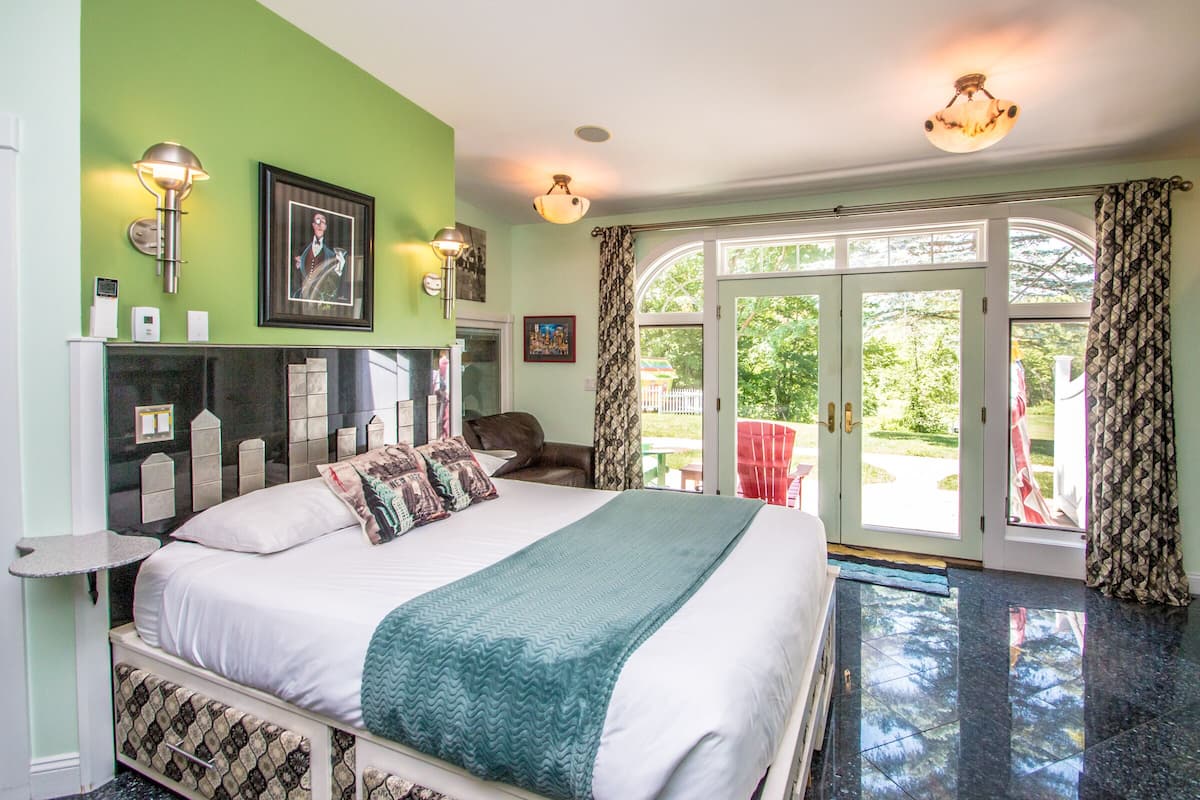 A bedroom with a bed, a nightstand, a lamp, a window with curtains, and a view of the outdoors.