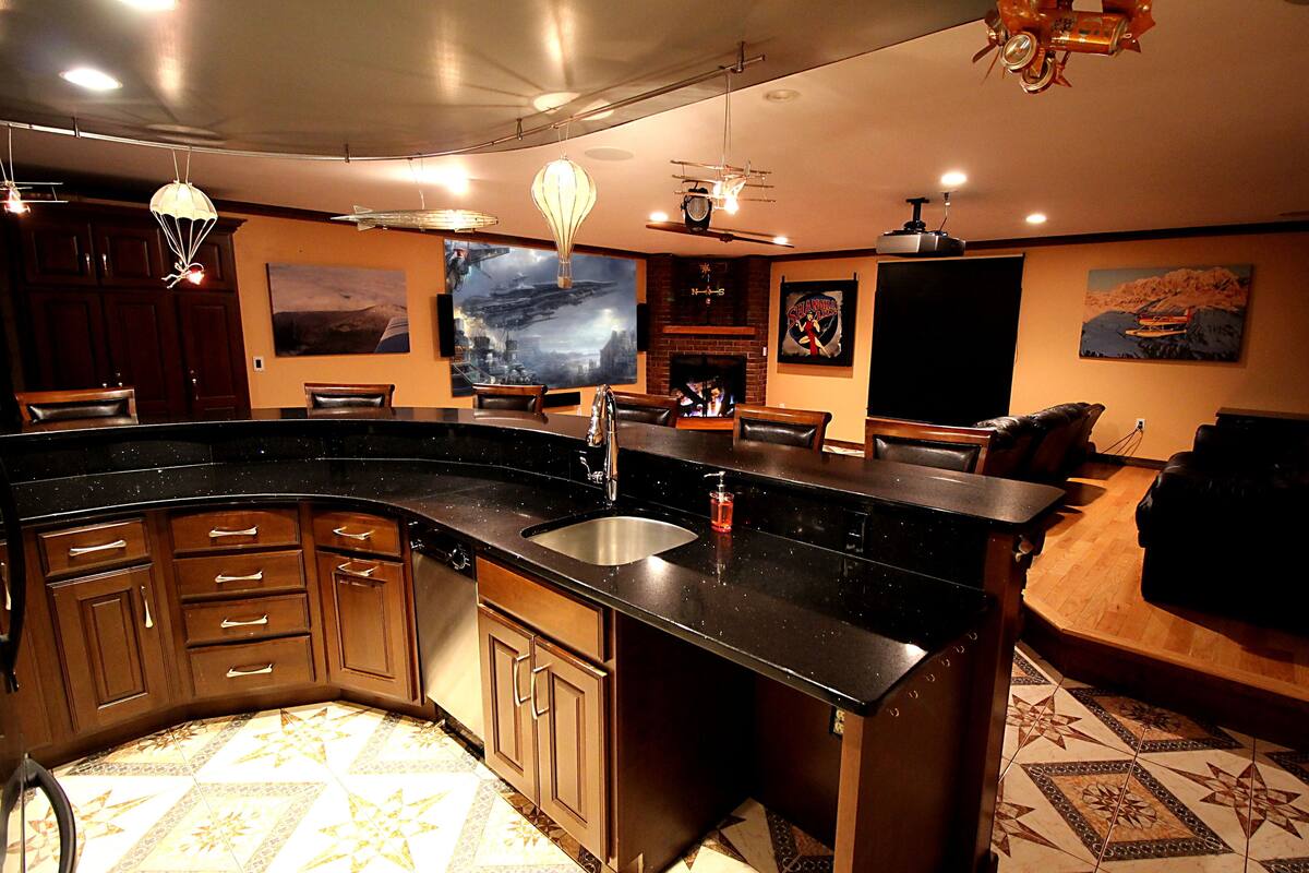 A modern kitchen with a curved island, dark countertops, and wooden cabinets. There is a large painting on the wall and a fireplace in the background.