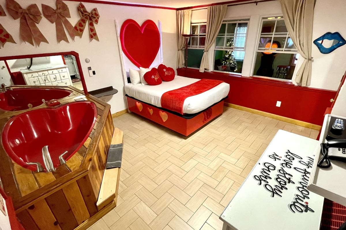 A bedroom with a large red heart decoration, a wooden bathtub, a bed with red bedding, and a mirror.