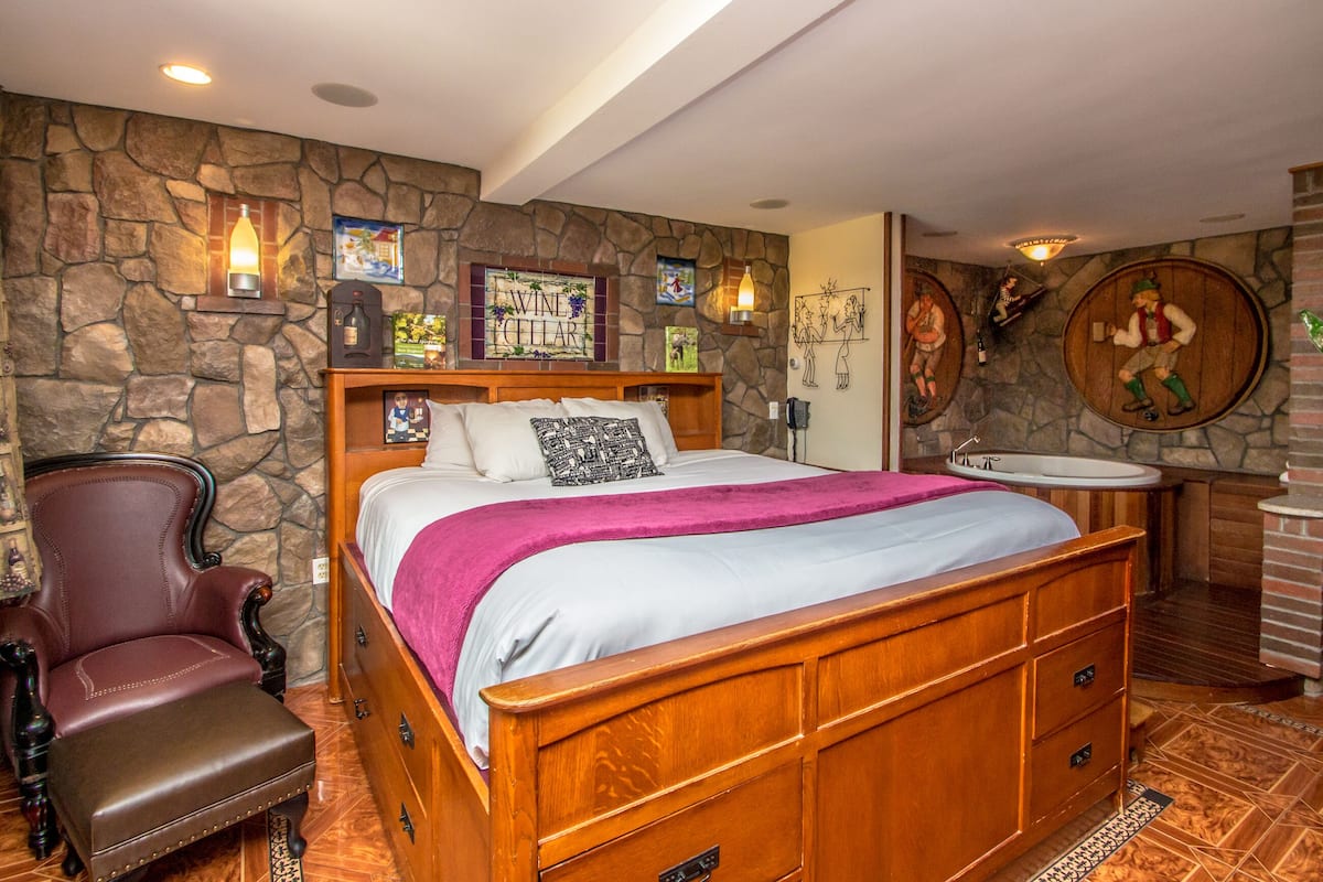 A bedroom with a stone wall, a wooden bed, a chair, and a bathroom with a sink and mirror.