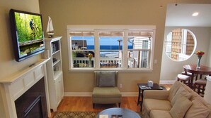 A living room with a fireplace, a television mounted above it, a sofa, a coffee table, and a view of the ocean through large windows. - Luxury Condo at the Arch in the heart of Historic Nye Beach (Newport)