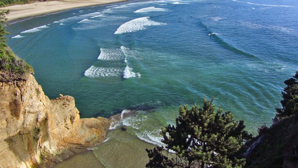 A coastal landscape with a beach, ocean waves, and forested hills. - Luxury Condo at the Arch in the heart of Historic Nye Beach (Newport)