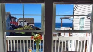 A window with a view of the ocean and houses, a table with a glass and flowers, and a chair. - Luxury Condo at the Arch in the heart of Historic Nye Beach (Newport)