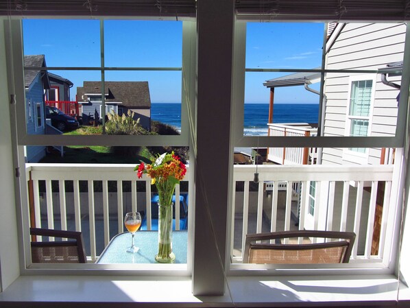 Una ventana con vista al océano y casas, una mesa con un vaso y flores, y una silla.
