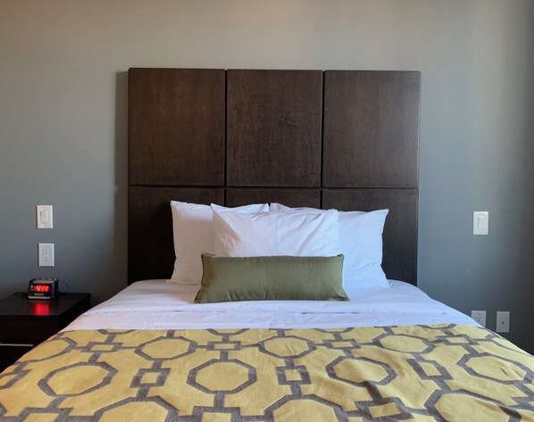 A hotel room with a bed, a nightstand with a clock, and a wall socket. - Voyage Hotel (Long Island City)