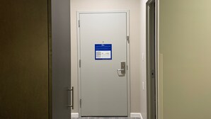 A hotel room with a wardrobe, a bed with a patterned bedspread, a door with an evacuation plan sign, and a light ceiling.