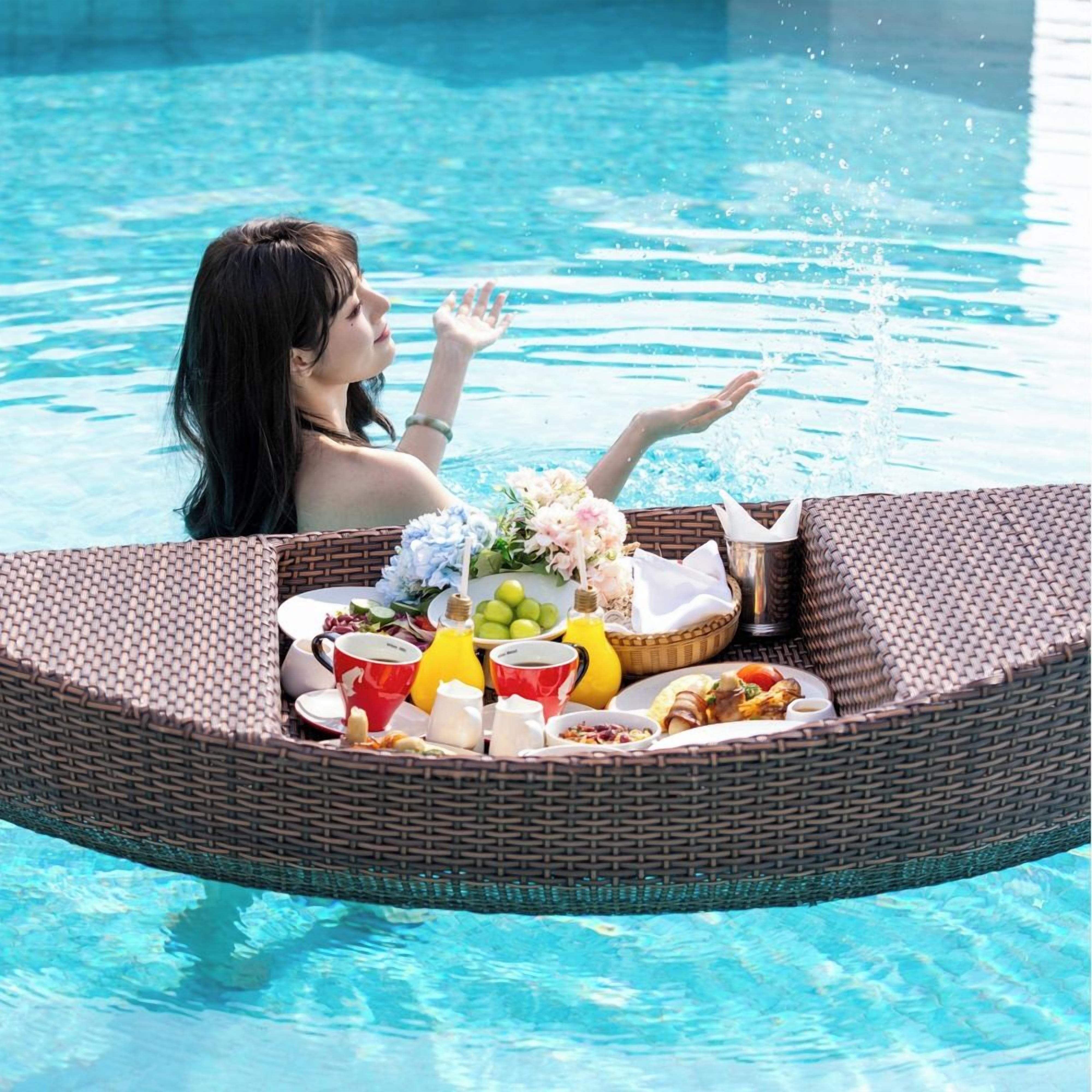 A person in a pool with a wicker lounge chair containing a meal and drinks.