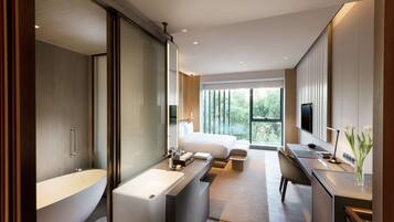 Une chambre d’hôtel moderne dotée d’une grande baignoire, d’une douche séparée, d’un lit avec vue, d’un bureau avec une chaise et d’une télévision.