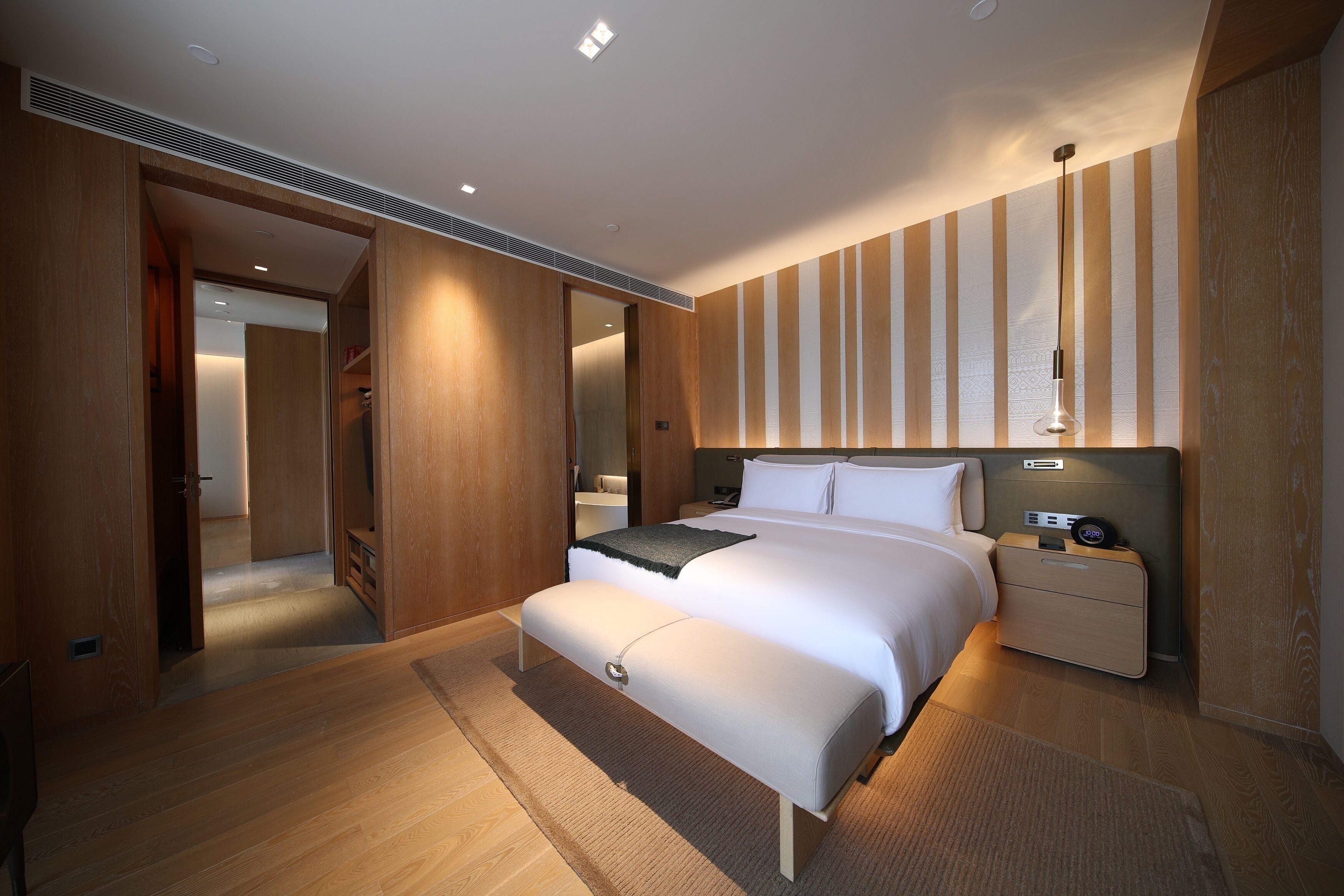 Una habitación de hotel moderna con una cama grande, mesitas de noche, un banco y una lámpara de pared.