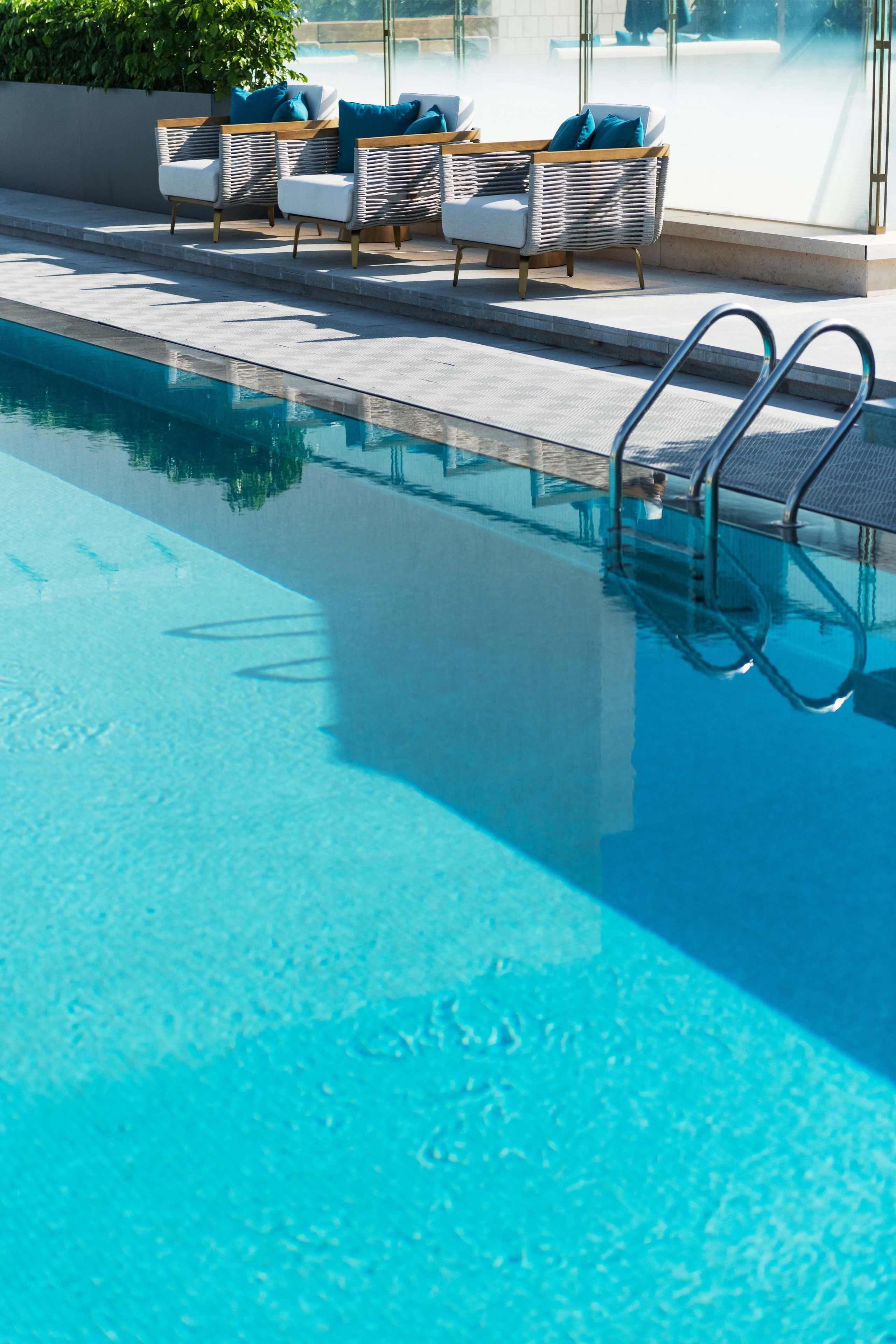 Una piscina con agua azul cristalina, una escalera metálica y sillones de mimbre con cojines.