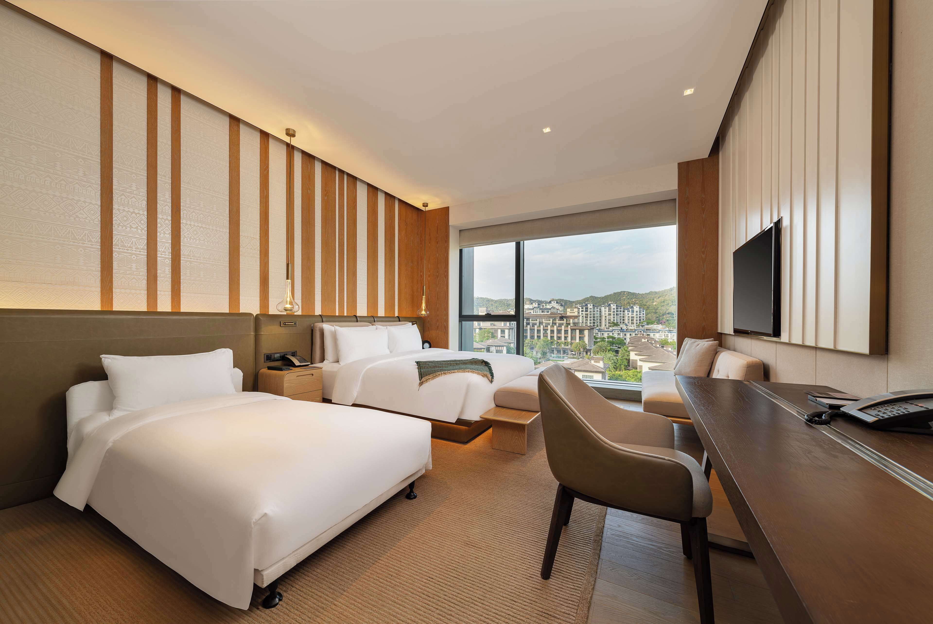 A hotel room with two beds, a desk, a chair, a TV, and a large window with a city view.