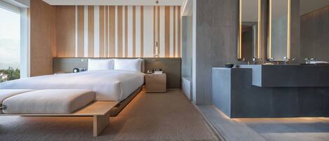 A modern hotel room with a large bed, bedside tables, a built-in bench, a large mirror, and a bathroom with a vanity and sink.