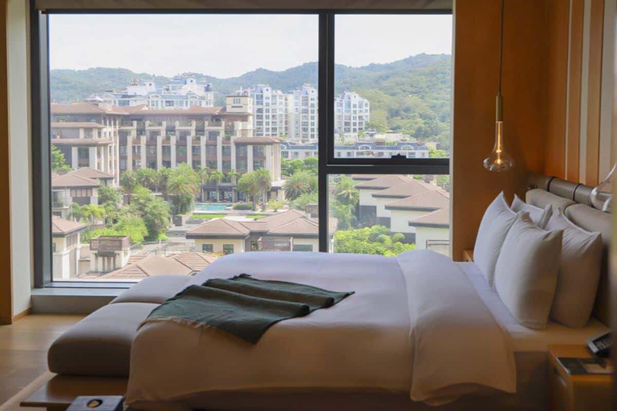 Une chambre d’hôtel avec un grand lit, une vue sur les bâtiments et la verdure, et un luminaire suspendu.