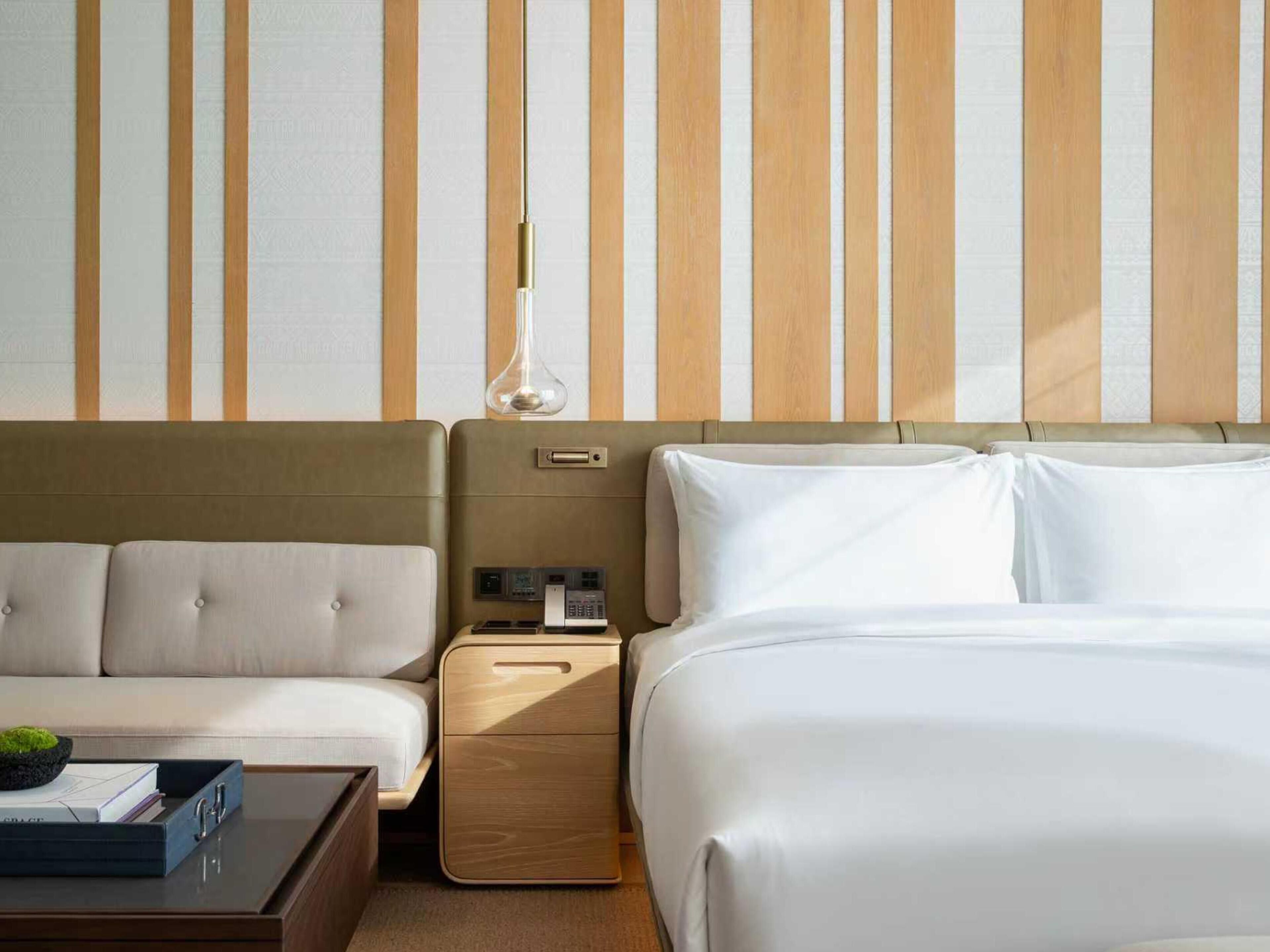 A hotel room with a striped wall, a bed, a sofa, a bedside table, and a lamp.