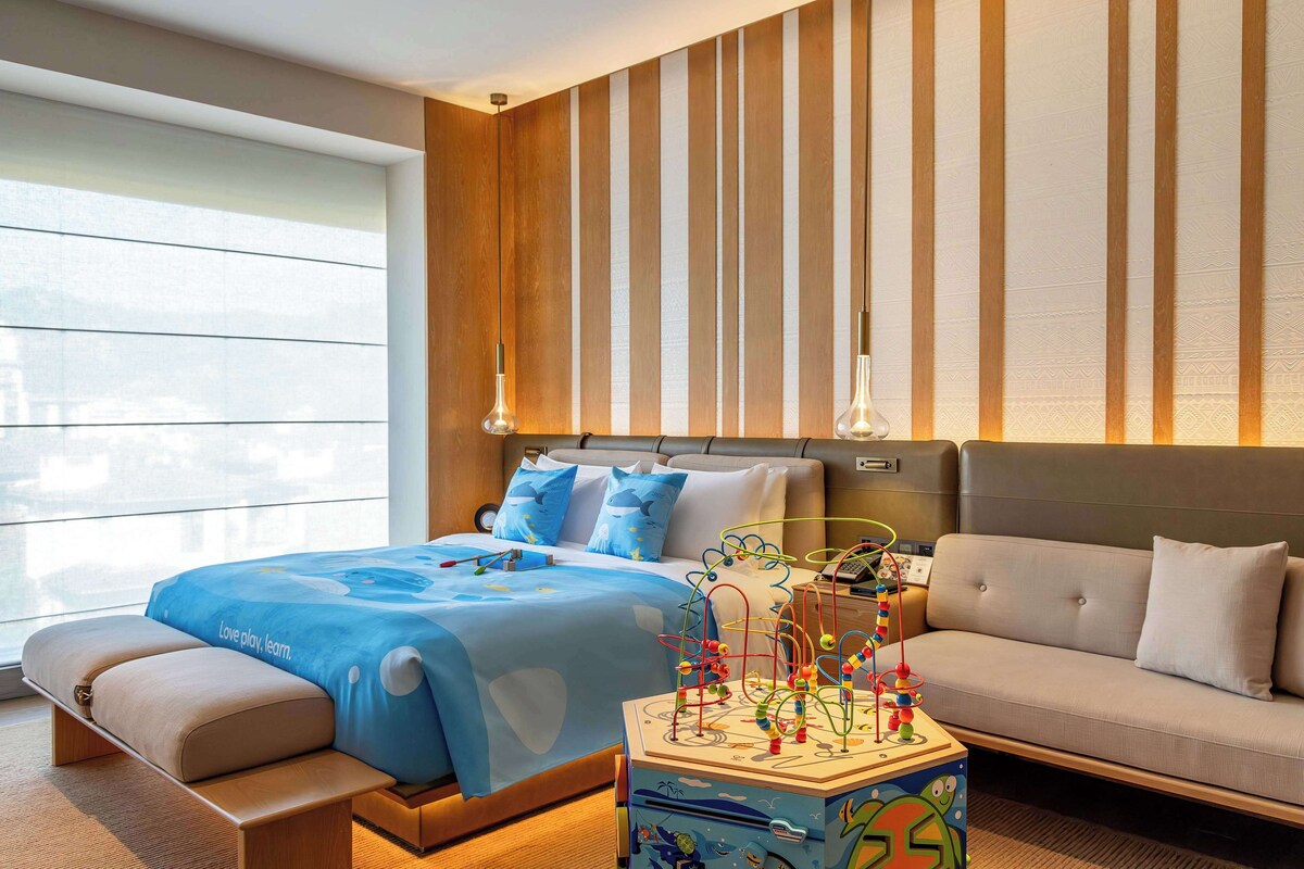 A modern hotel room with a large bed, a sofa, a play area with toys, and a large window.