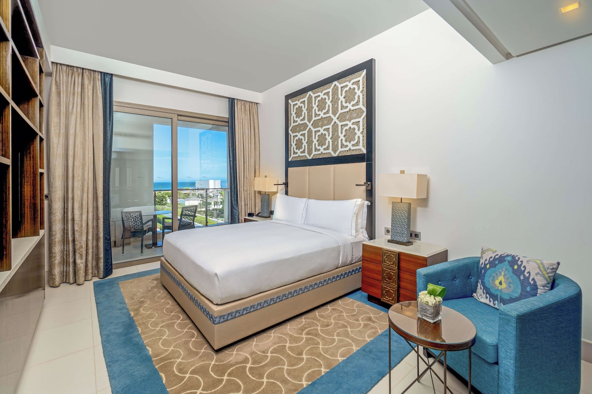 Quarto de hotel com uma cama grande, um sofá azul, uma mesa pequena e vista para a cidade através de uma janela ampla.
