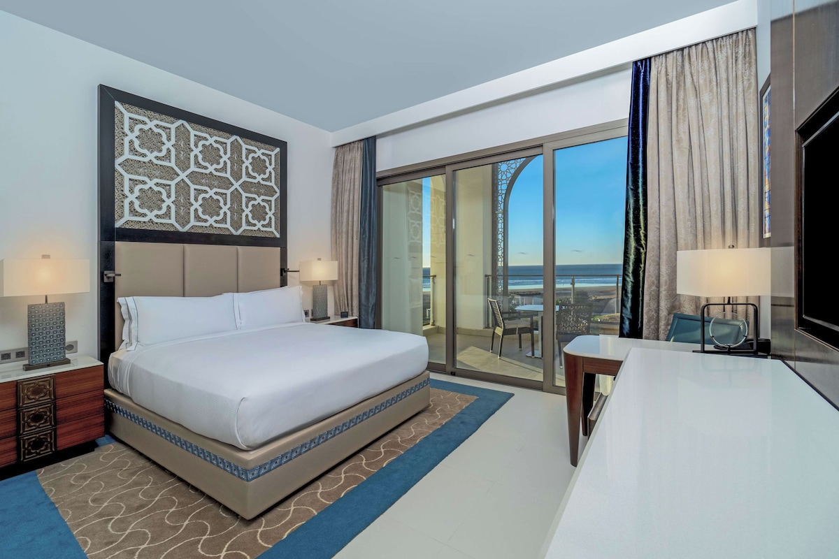 Quarto de hotel com uma cama grande, uma mesa, uma cadeira e vista para o mar através das portas de vidro.