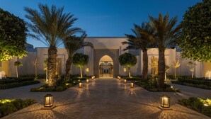 A well-lit pathway leading to a building entrance, flanked by palm trees and lit lanterns.