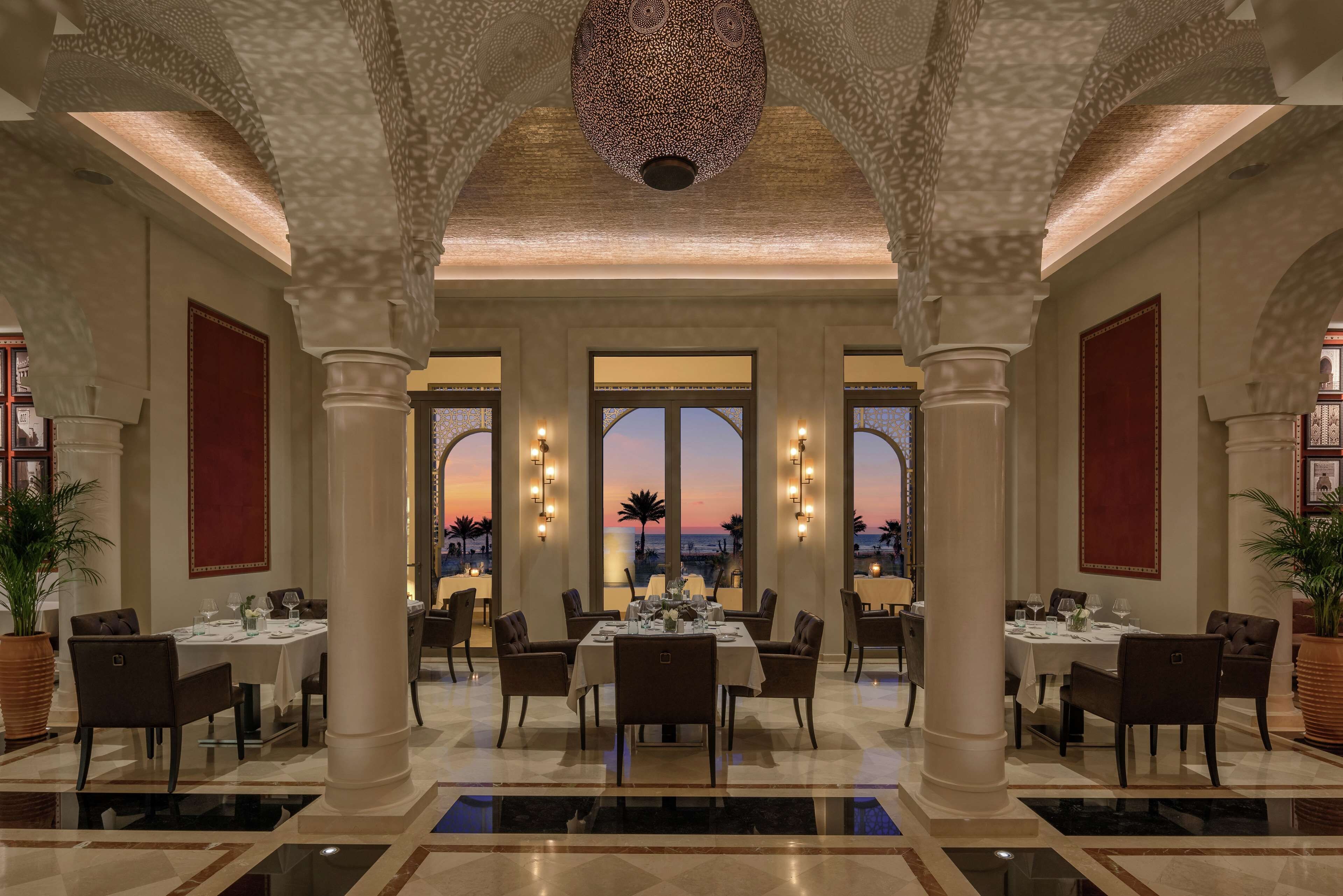A spacious dining area with high ceilings, large windows, and a view of the sunset.