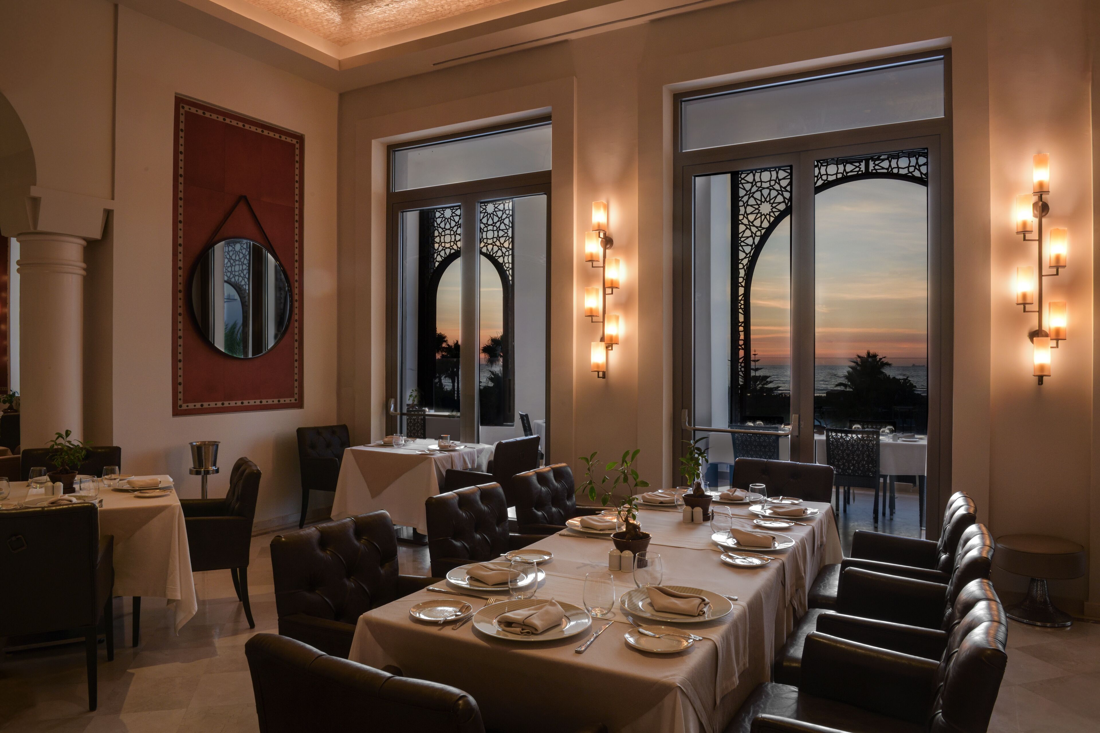 Comedor con mesas dispuestas para una comida, ventanas amplias con paneles decorativos e iluminación empotrada.