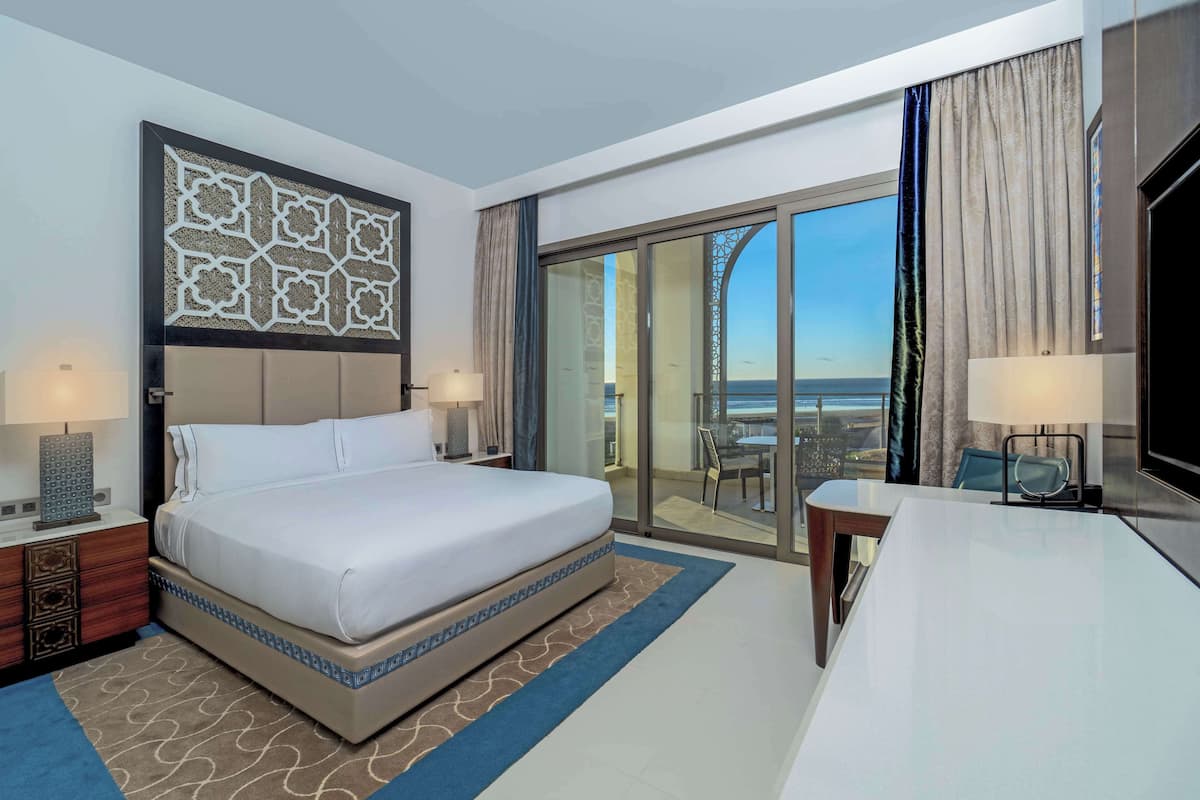Une chambre d’hôtel avec un grand lit, un bureau, une chaise et une vue sur la mer, visible à travers les portes vitrées.