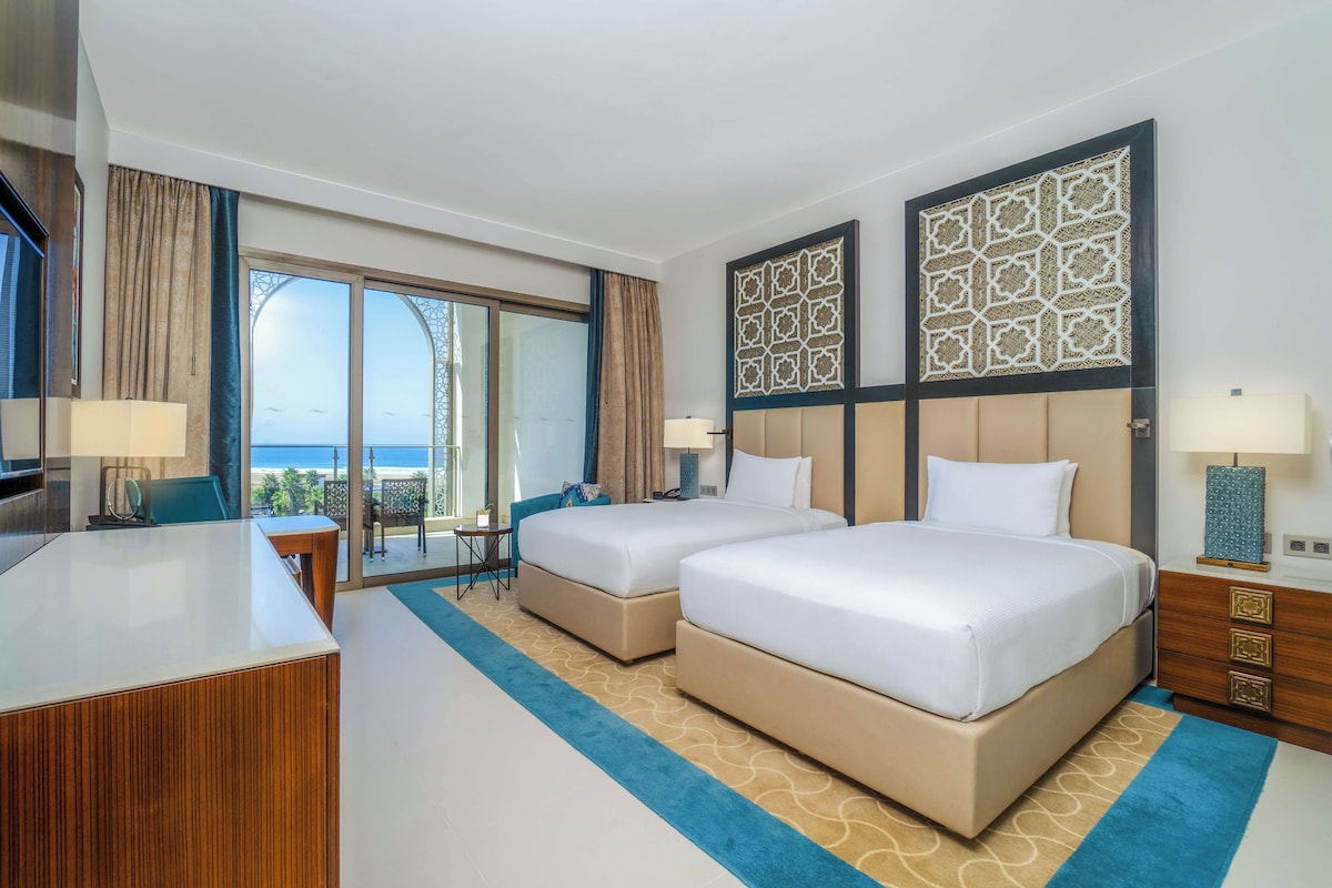 Quarto de hotel com duas camas, uma escrivaninha, uma televisão e vista para o exterior.