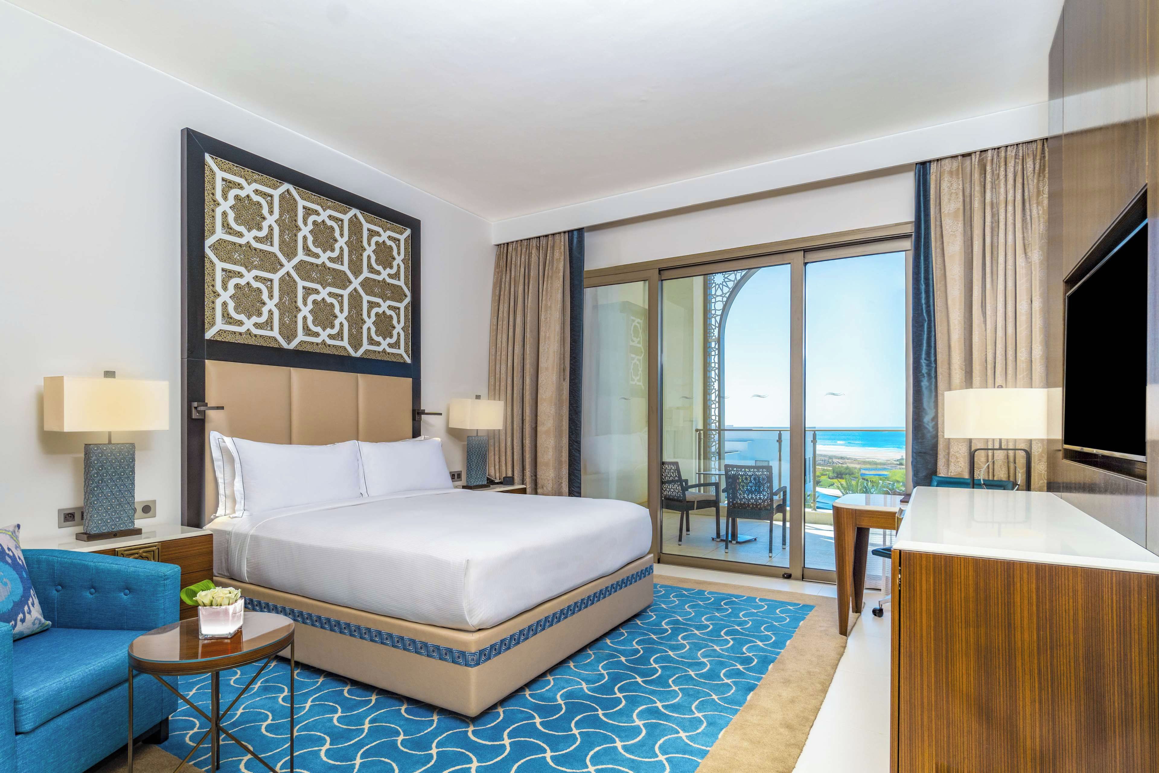 Ein Hotelzimmer mit einem großen Bett, einem Schreibtisch, einem Sessel, einem Fernseher und einem Balkon mit Blick aufs Meer.