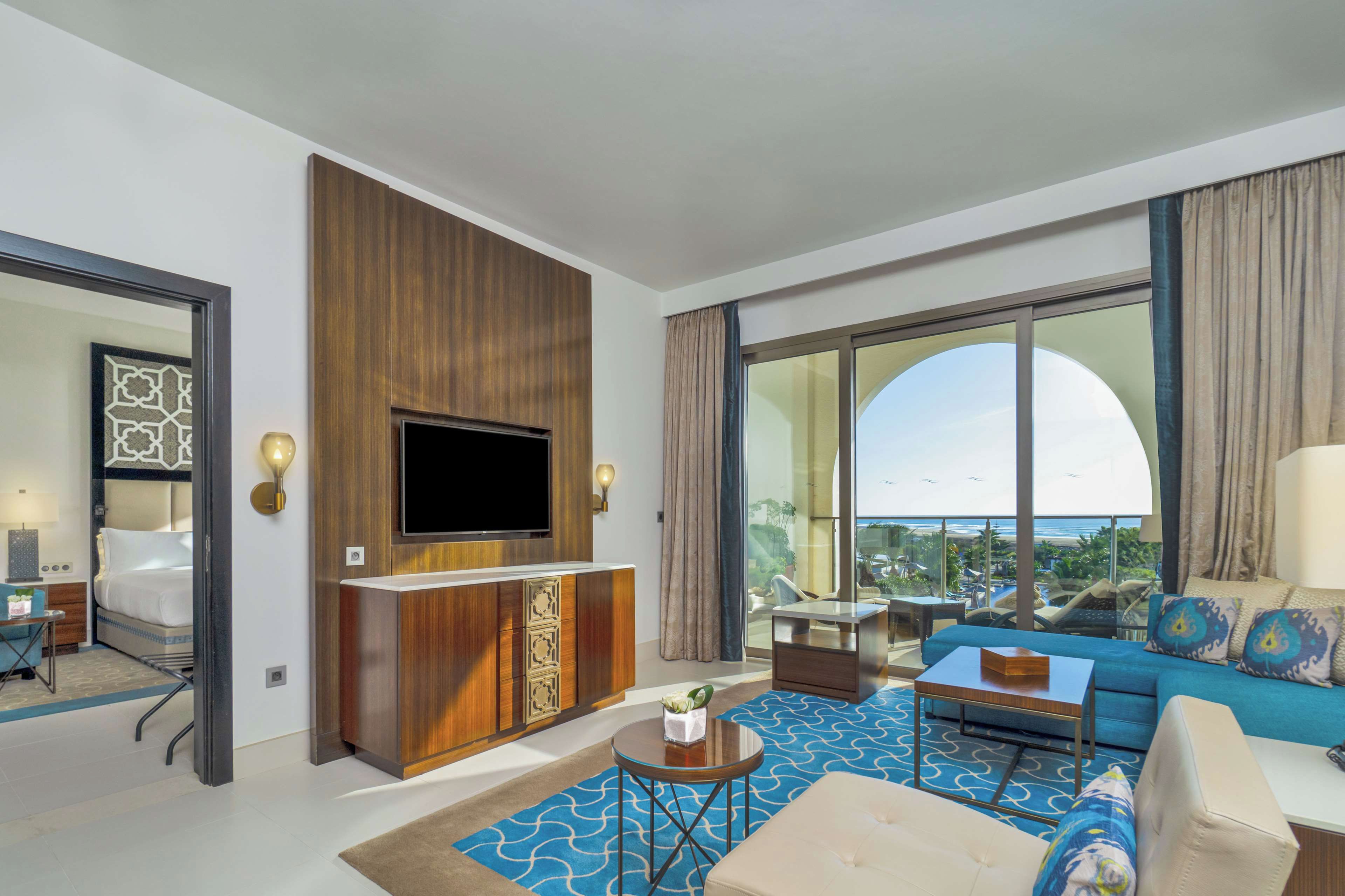 A hotel room with a large TV, a sofa, a coffee table, and a view of the ocean.