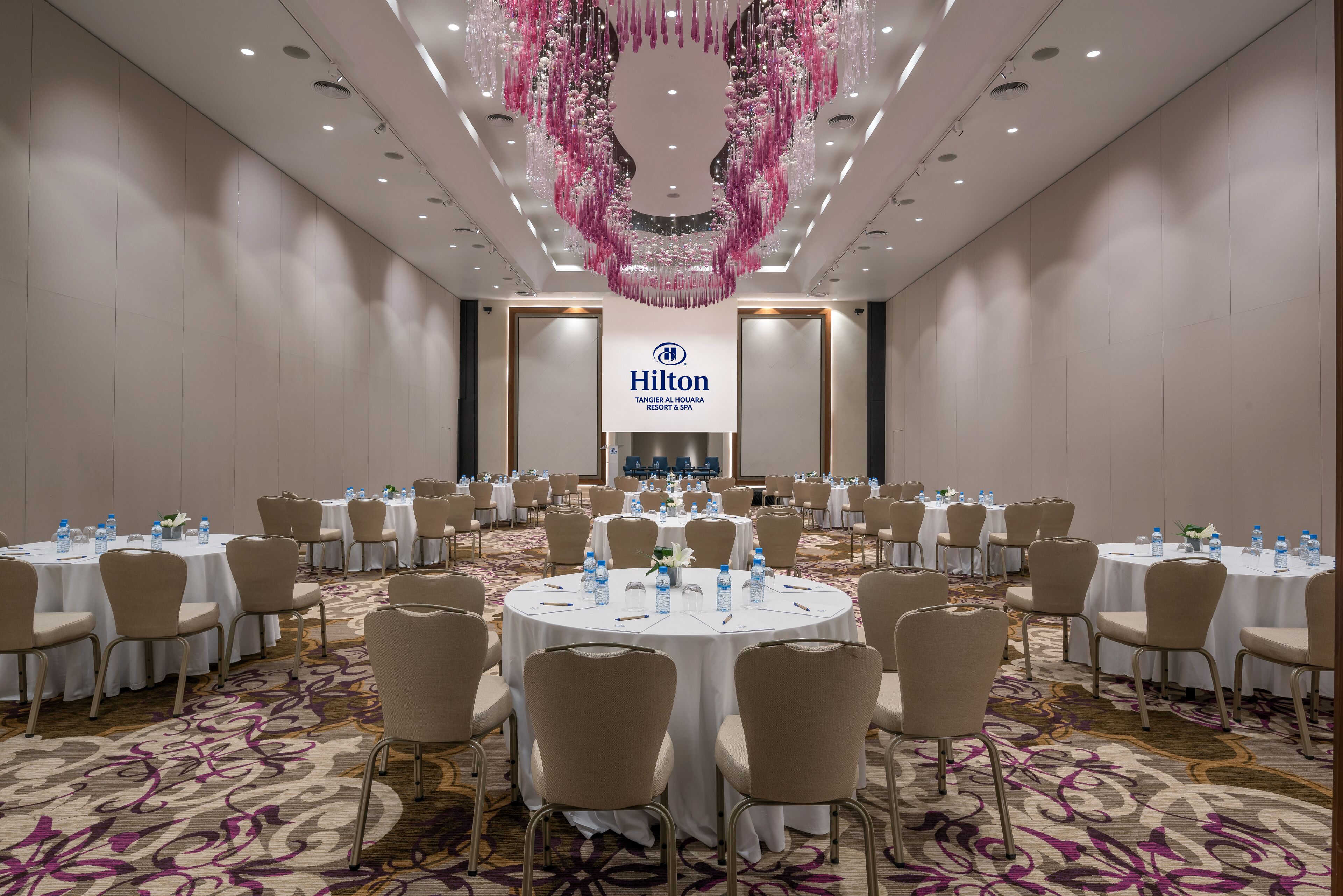 A large conference room with round tables, beige chairs, and a floral carpet. A Hilton logo is visible on the wall.