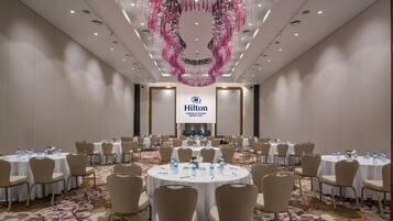 A large conference room with round tables, beige chairs, and a floral carpet. A Hilton logo is visible on the wall.