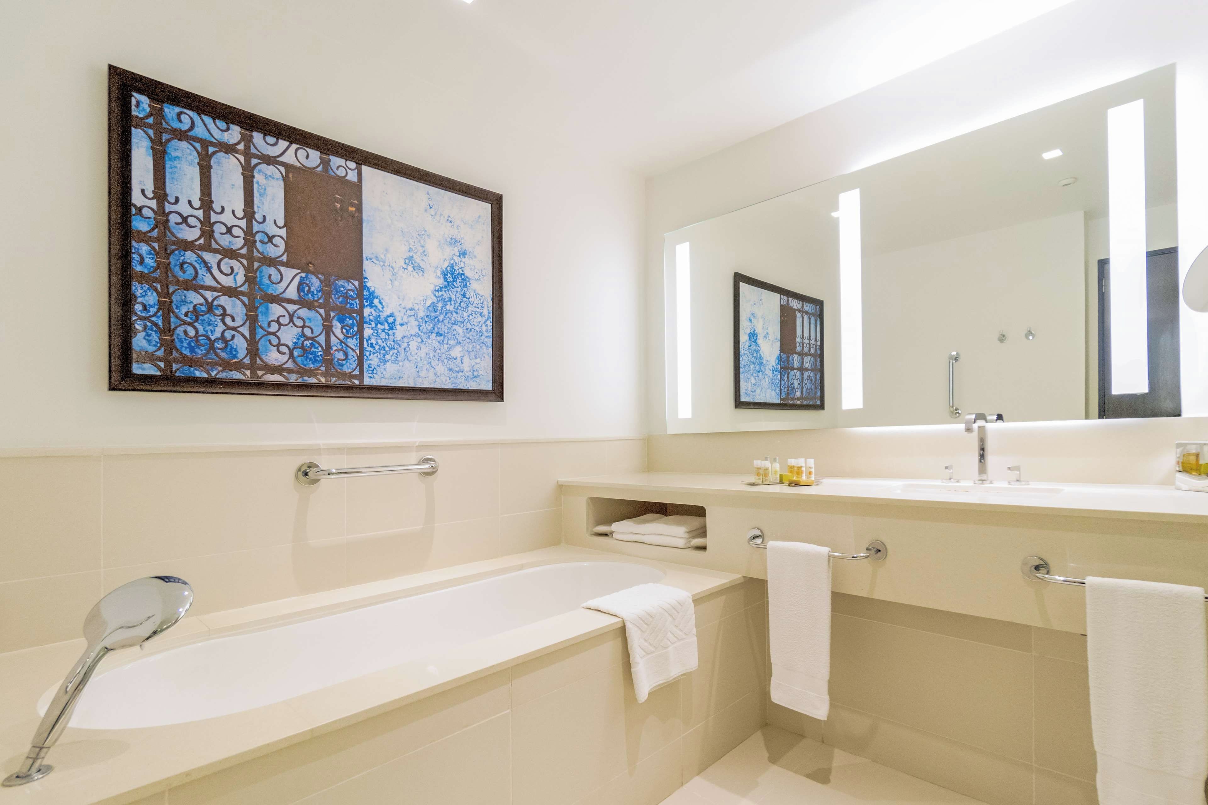 A modern bathroom with a bathtub, a large mirror, and a framed artwork on the wall.