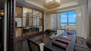 A modern hotel room with a large window offering a view of the beach, a dining area with a long table, and a cozy seating area with a sofa and chairs.