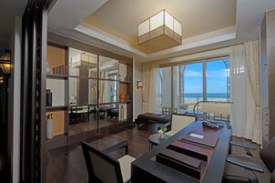 Una habitación de hotel moderna con un ventanal grande que ofrece vistas a la playa, un comedor con una mesa larga y un rincón de descanso acogedor con un sofá y sillones.