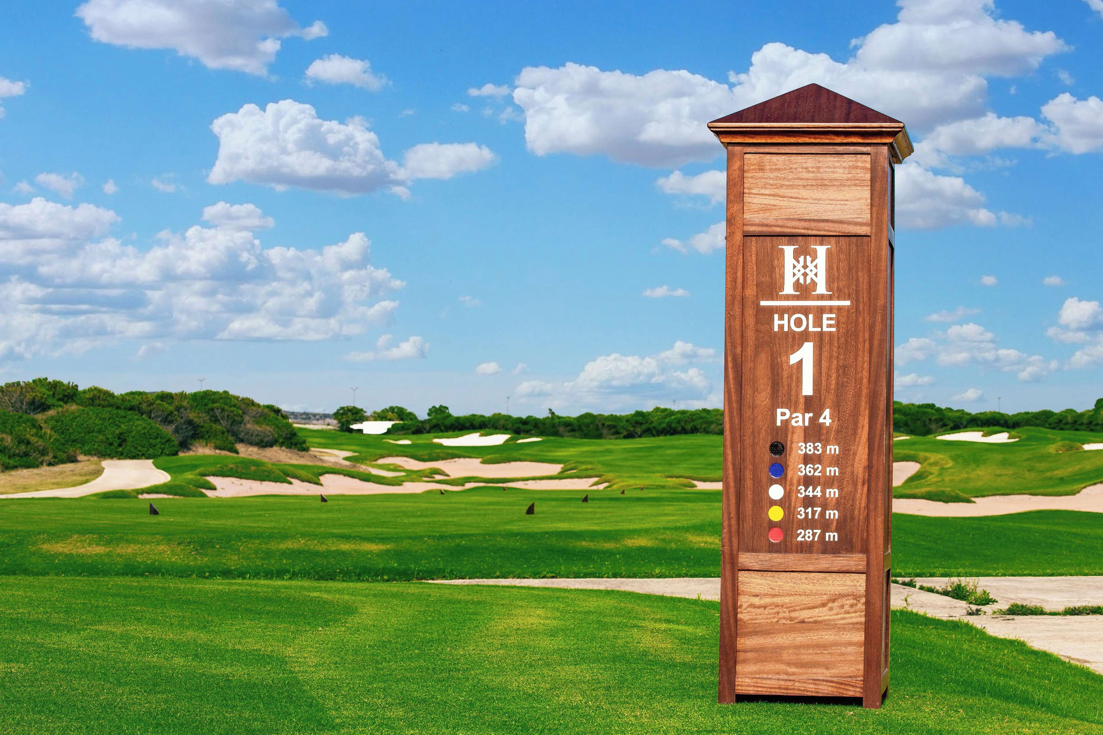 Placa de madeira indica 'HOLE 1' e 'Par 4', marcando um buraco de golfe em um campo.