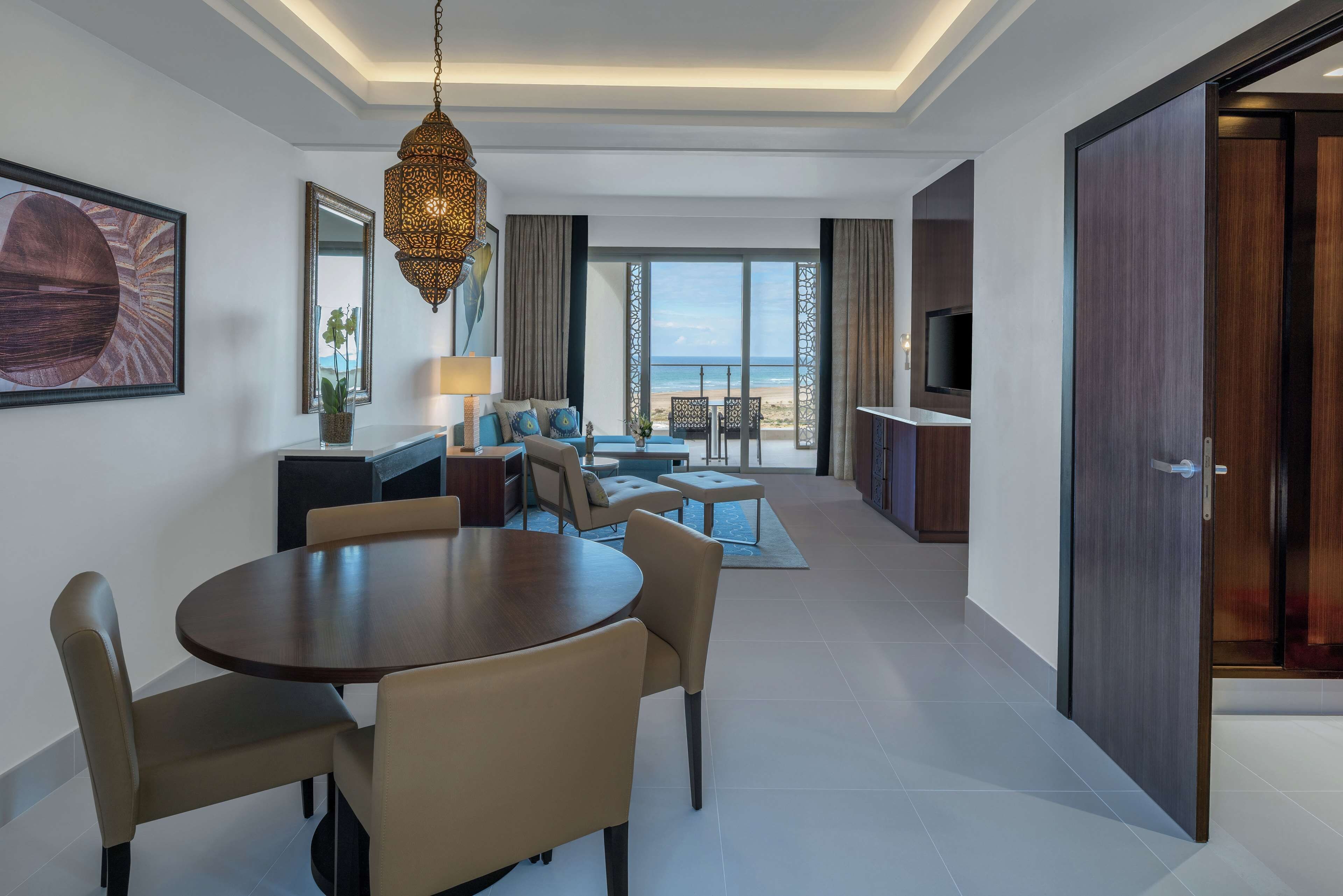 A modern hotel room with a dining area, a TV, and a view of the beach.