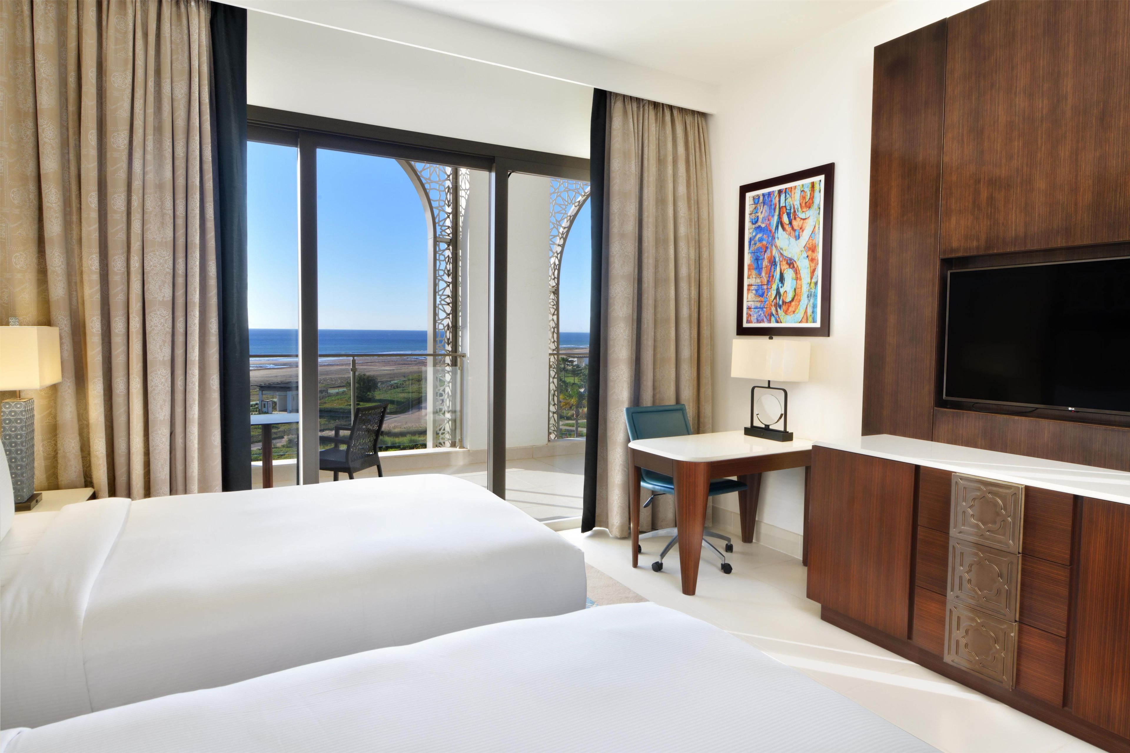 A hotel room with two beds, a desk, a chair, a TV, and a large window with a view of the sea.