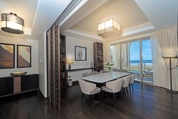 A modern dining area with a large table and chairs, surrounded by bookshelves and a view of the ocean.