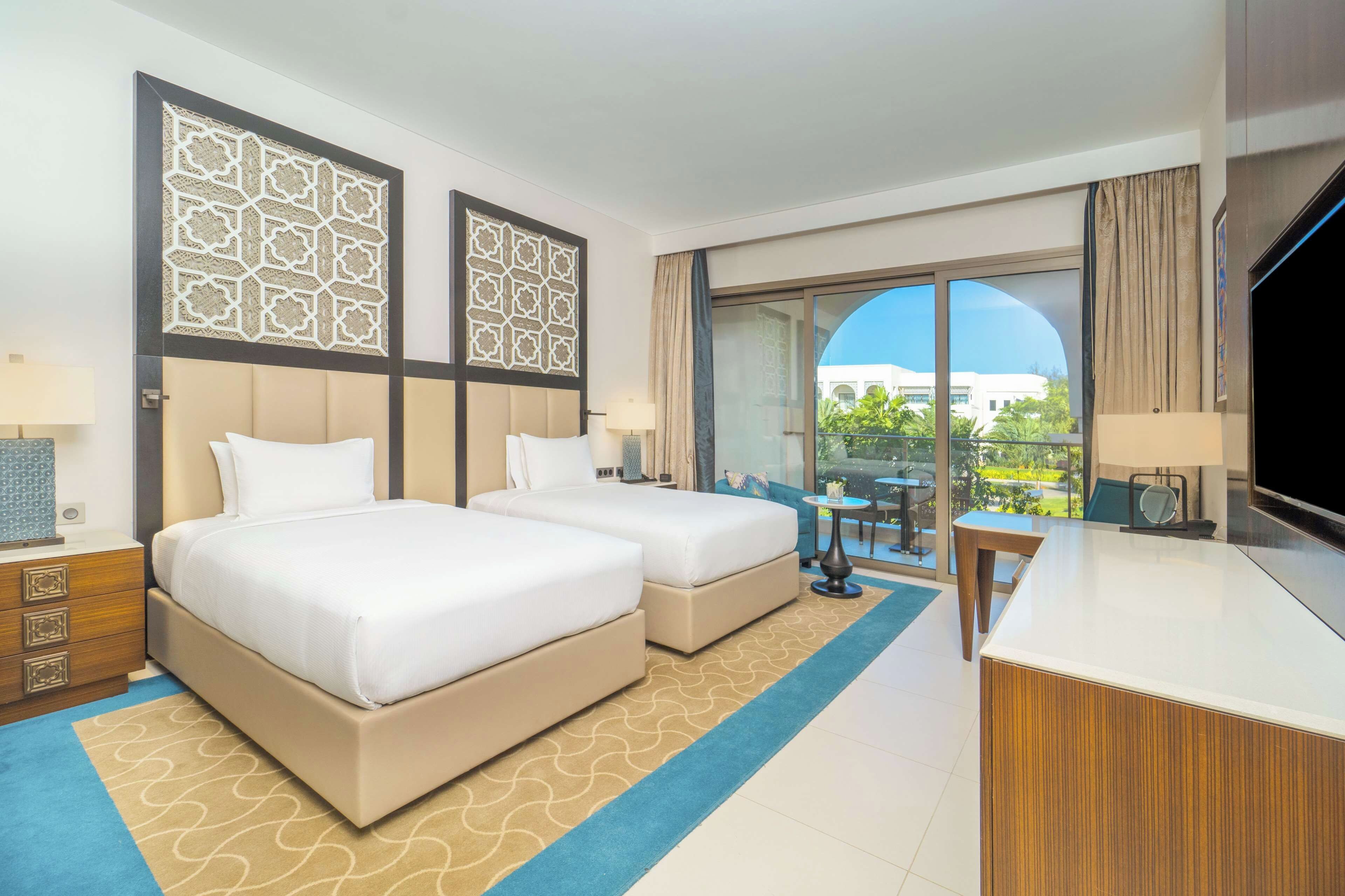 A hotel room with two beds, a desk, a TV, and a balcony with a view of the city.