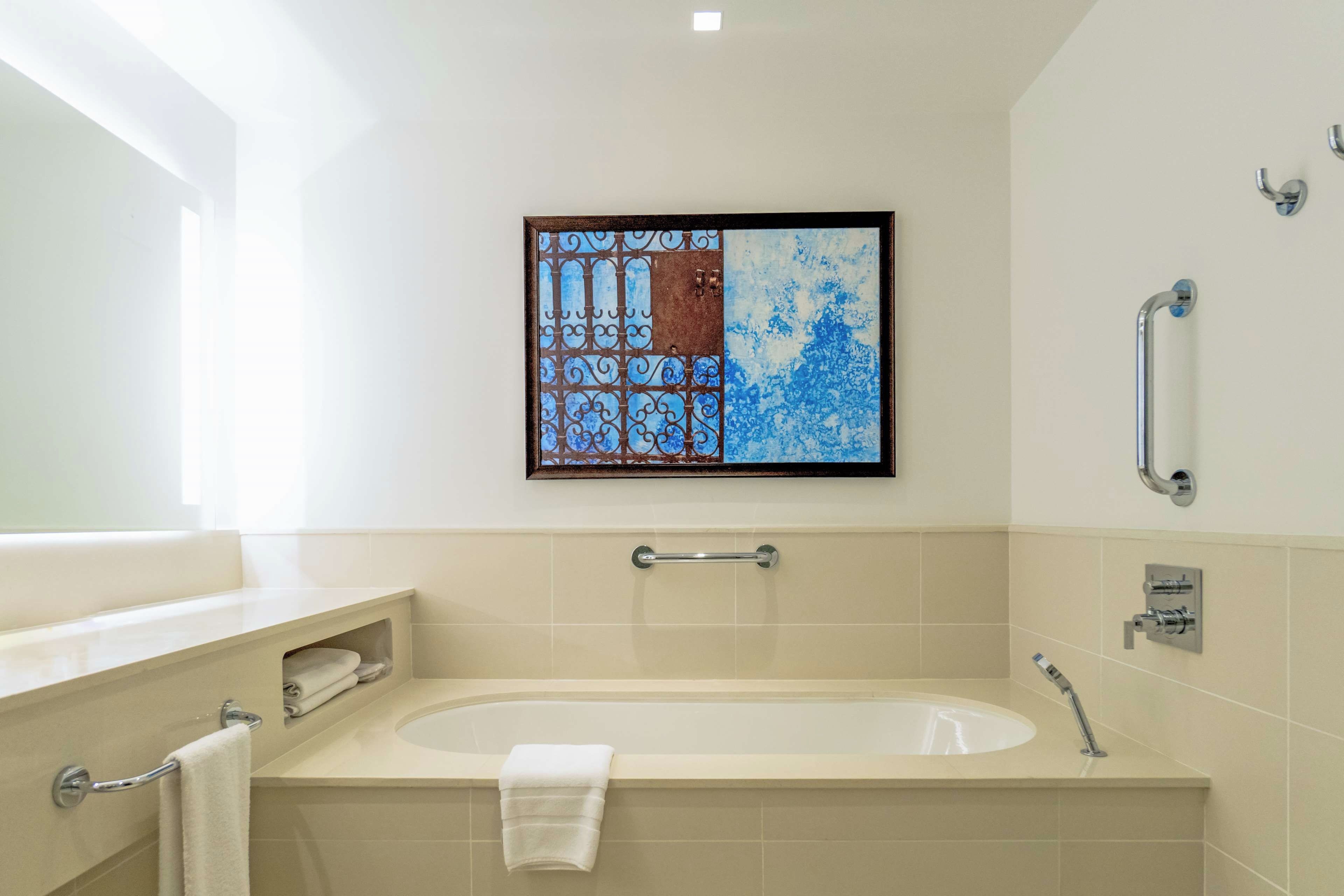 A modern bathroom with a bathtub, a towel rack, a handrail, and a framed artwork on the wall.