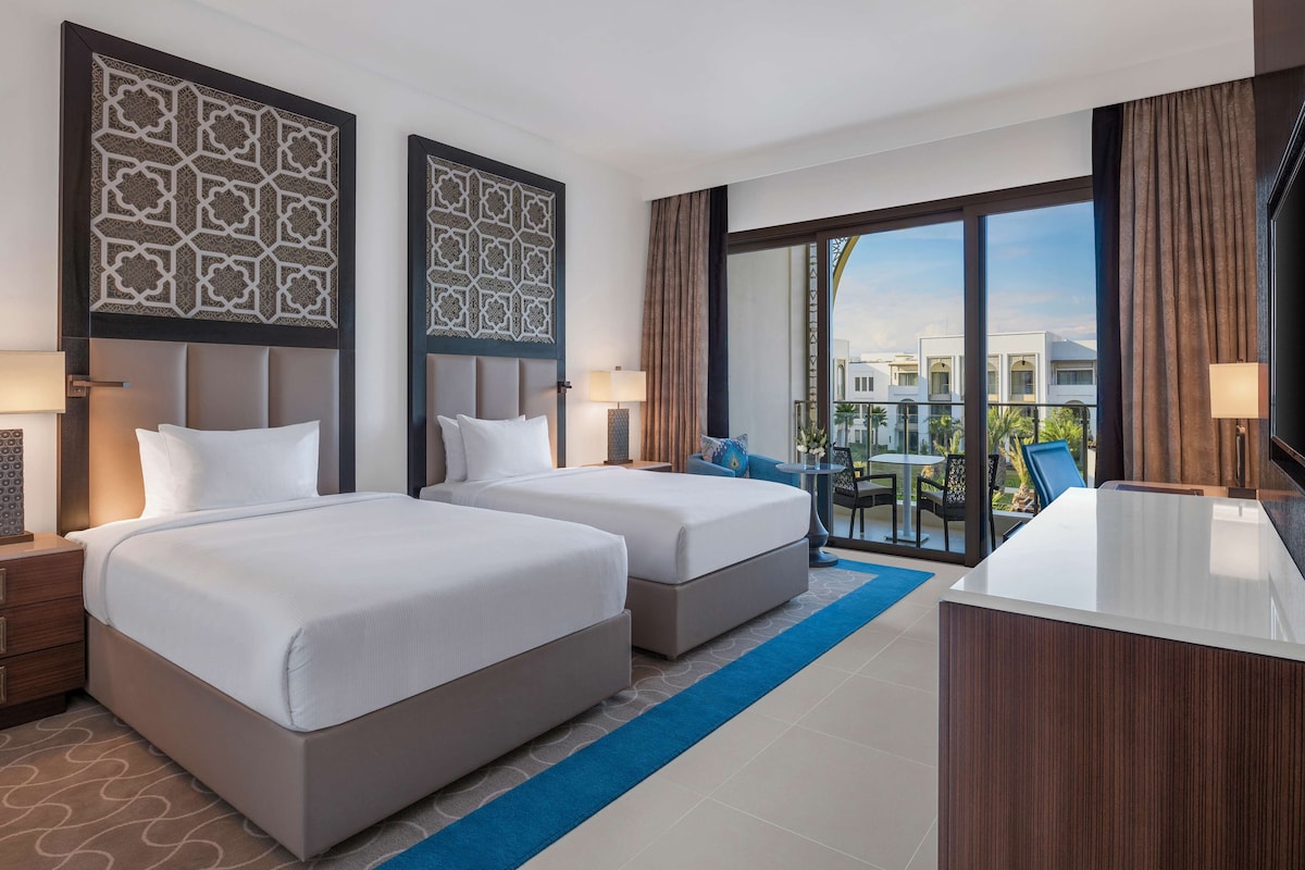 Quarto de hotel com duas camas, uma escrivaninha e varanda com vista para edifícios.